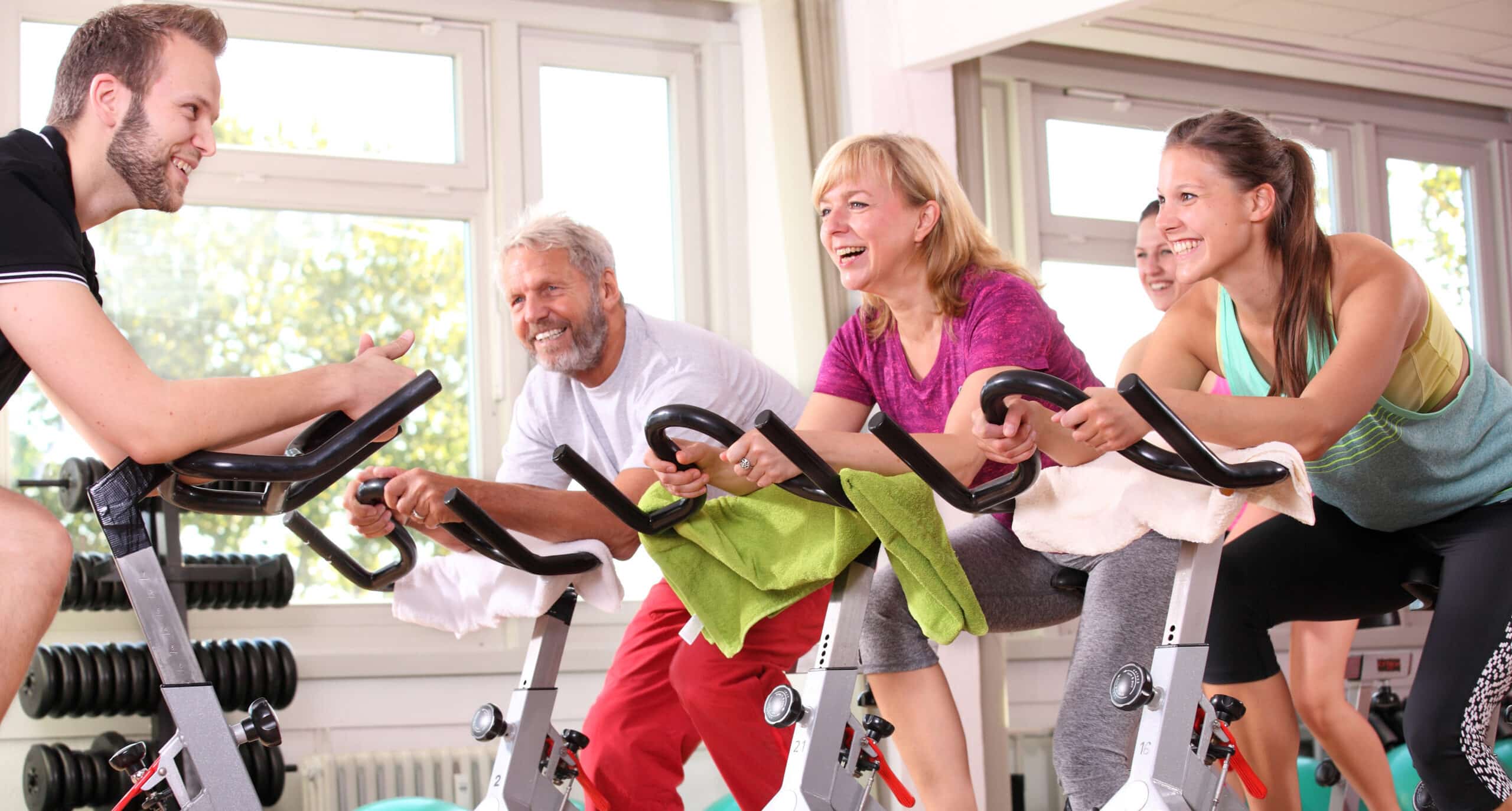 Ein Mann und zwei Frauen sitzen auf Spinningbikes, ihnen gegenüber sitzt Ihr Trainer ebenfalls auf einem Spinningbike.