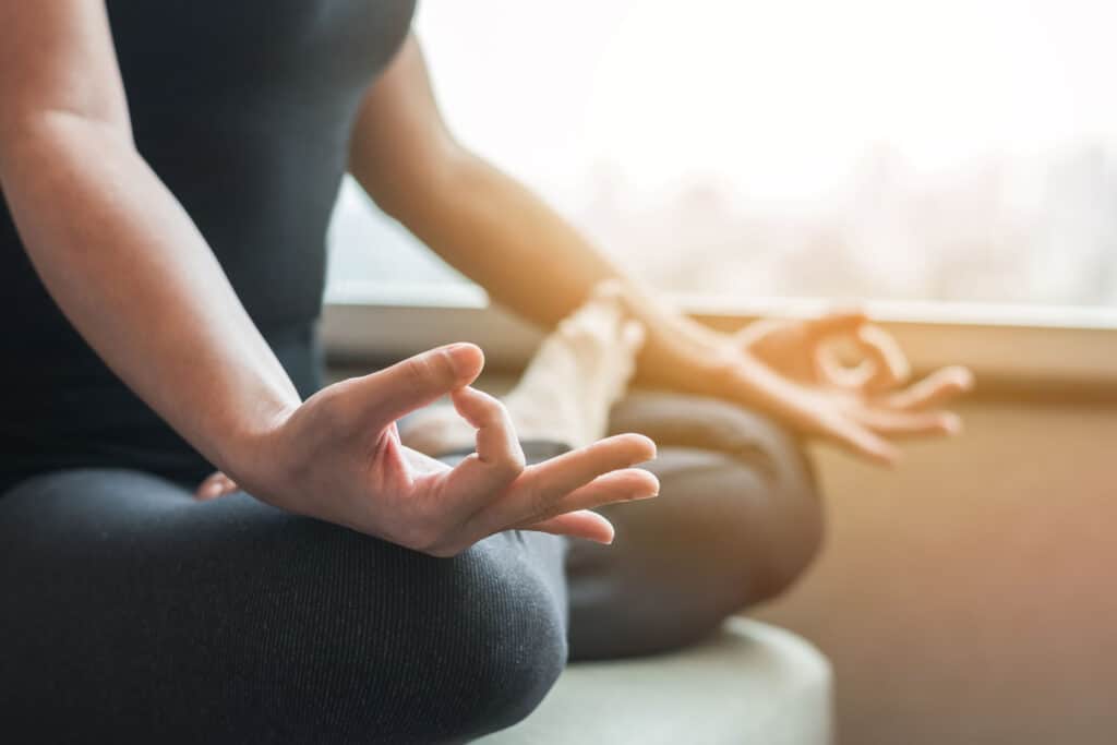 Eine Frau in schwarzem Sportoutfit sitzt im Schneidersitz auf dem Boden, ihre Zeigefinder und Daumen berühren sich und die Hände liegen auf ihren Knien.