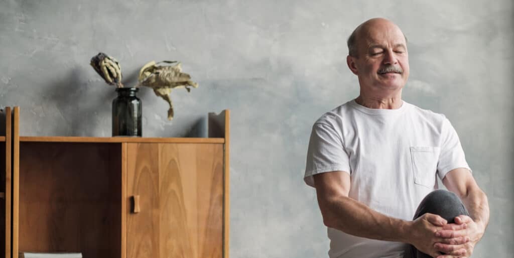 Ein älterer Mann mit Glatze und einem Schnurrbart steht auf einem Bein und meditiert. I mHIntergund sieht man eine graue Wand vor der ein modernes Holzregal steht.