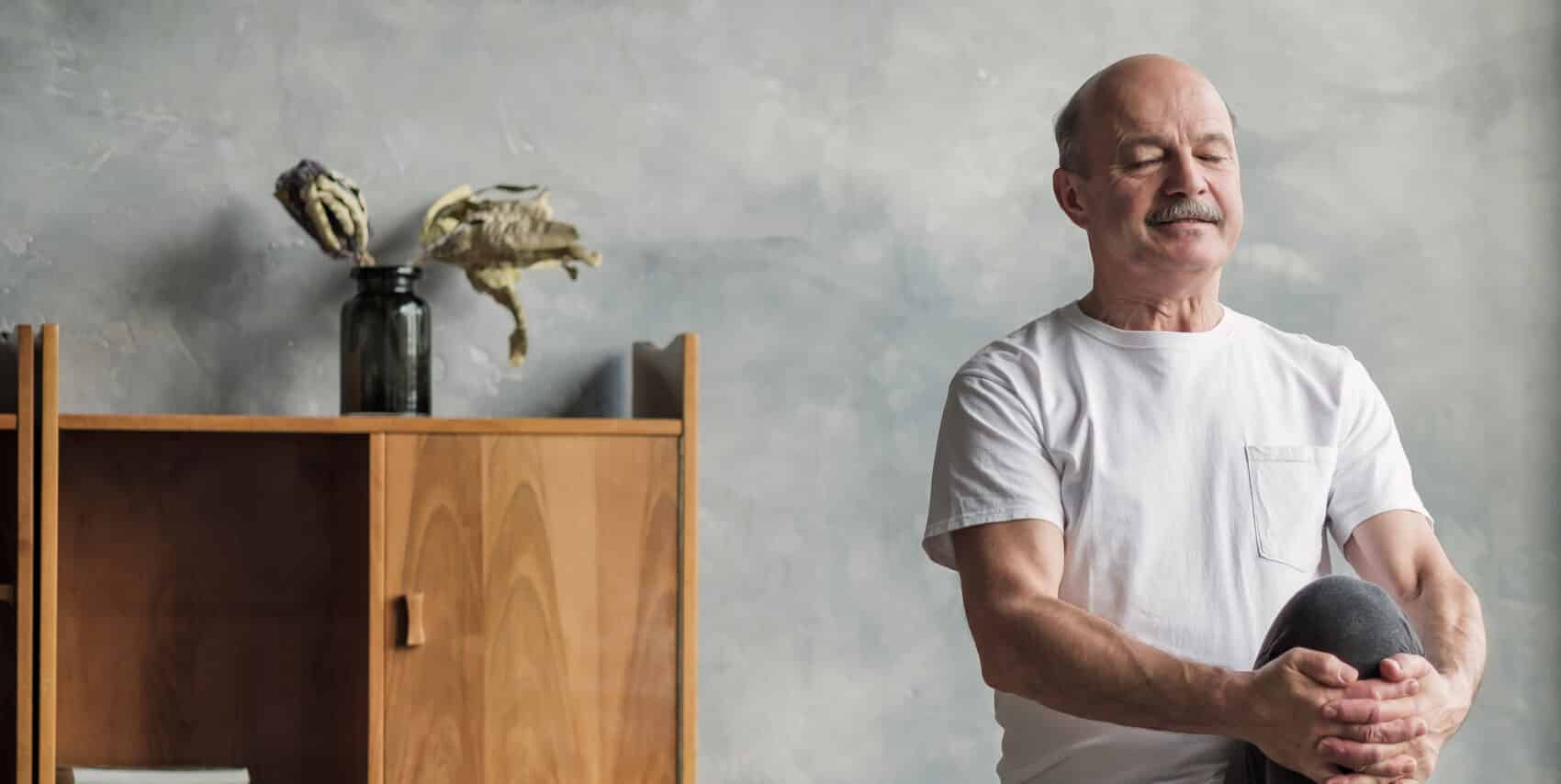 Ein älterer Mann mit Glatze und einem Schnurrbart steht auf einem Bein und meditiert. I mHIntergund sieht man eine graue Wand vor der ein modernes Holzregal steht.