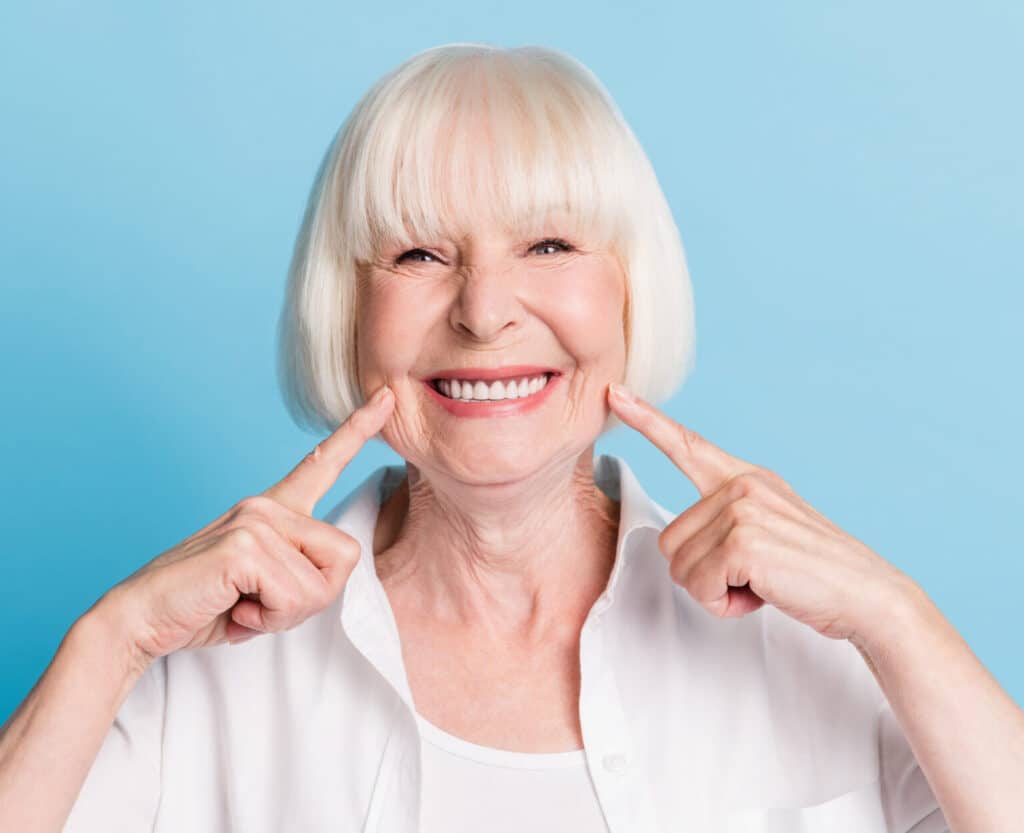 Eine ältere Frau mit weißem Bob grinst in die Kamera und drückt mit ihnen Zeigefingern in ihre Backen. Der Hintergrund ist Blau.