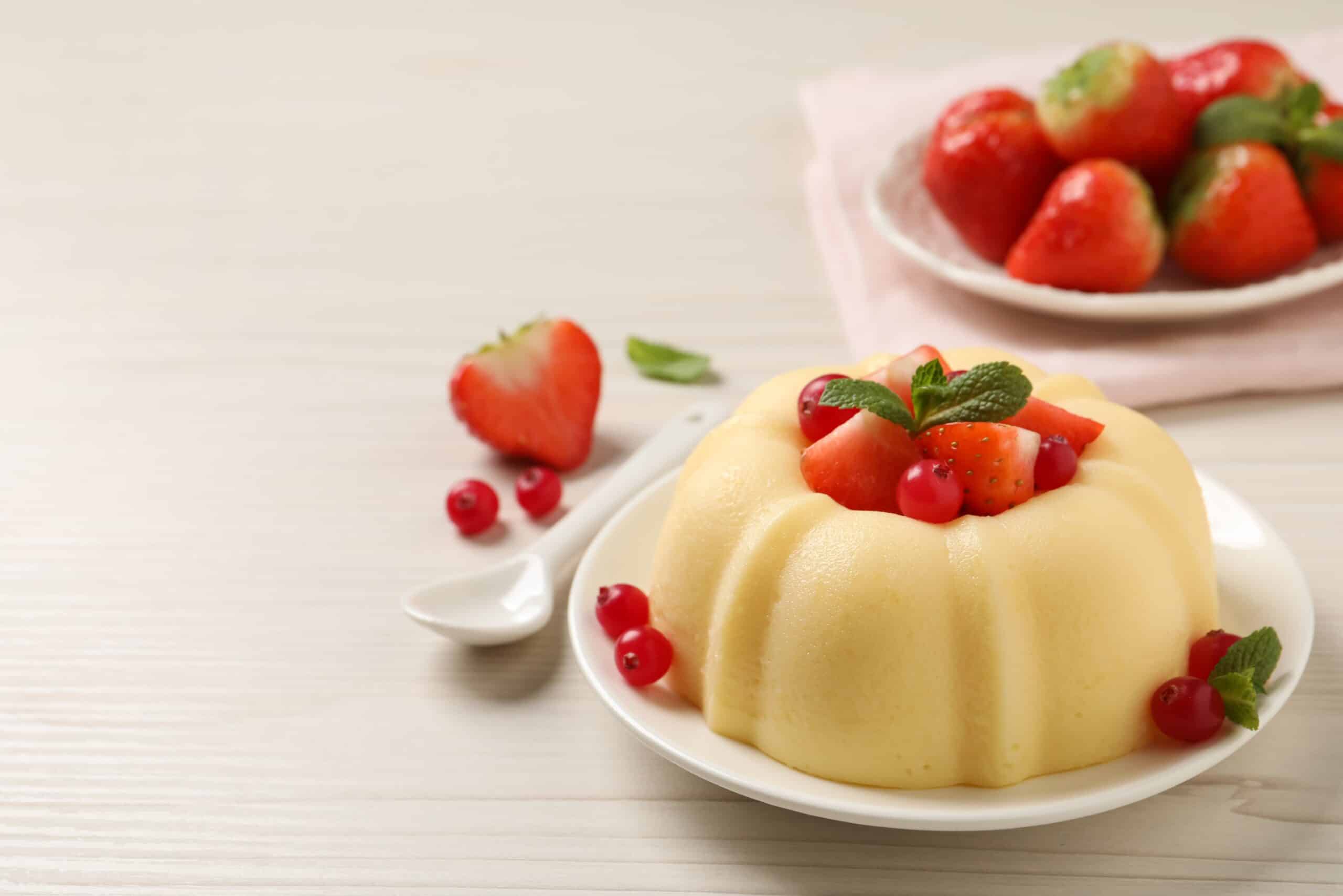 Gestürztes Buttermilchgelee/Pudding auf einem kleinen Teller, garniert mit frischen Beeren, neben einem weißen Löffel und vor einem Teller Erdbeeren auf hellem Tuch.